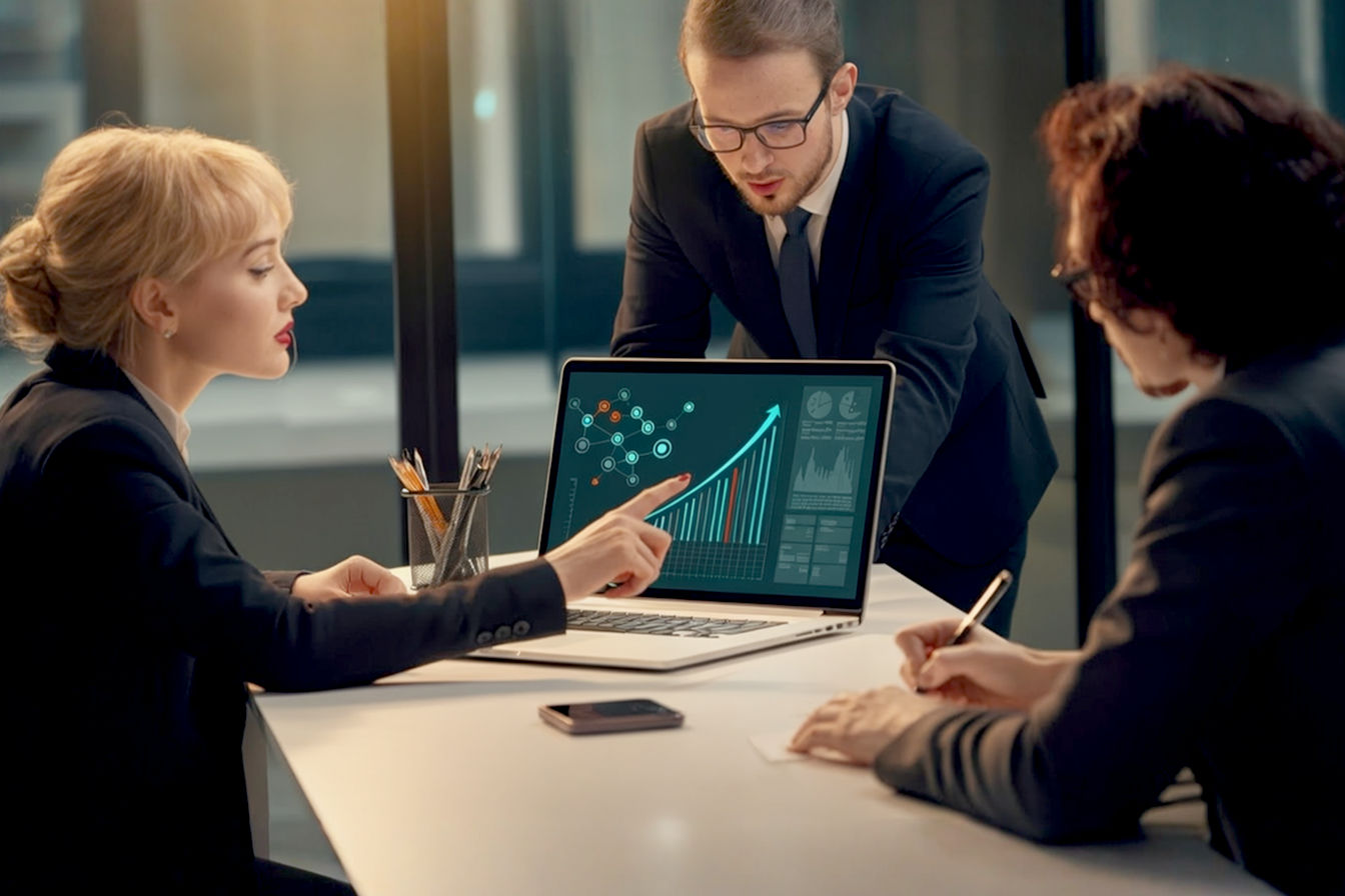 Professional, lifelike, eye-level medium shot photo. A team of three B2B professionals collaboratively working around a modern, minimalist office table. One person is pointing at a sleek laptop screen displaying abstract representations of growth charts and network connections (subtle, not overpowering). Another is taking notes, and the third is actively engaged in discussion, smiling. The atmosphere is bright, optimistic, and focused. Soft, natural light from a nearby window.