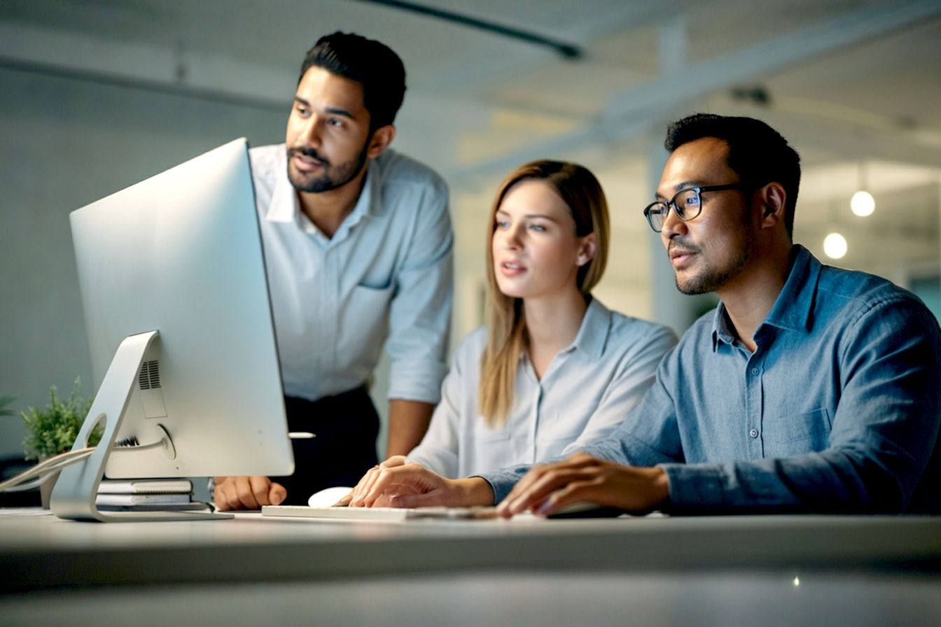 Professional, lifelike corporate photography style. A team of three business professionals are discussing search engine optimisation in front of a computer. The background is a bright, modern office interior with soft, natural lighting. The mood is optimistic, focused, and innovative. Emphasize clarity, a sense of connection, and growth. The computer screen should be the focal point, subtly hinting at digital strategy and data analysis.