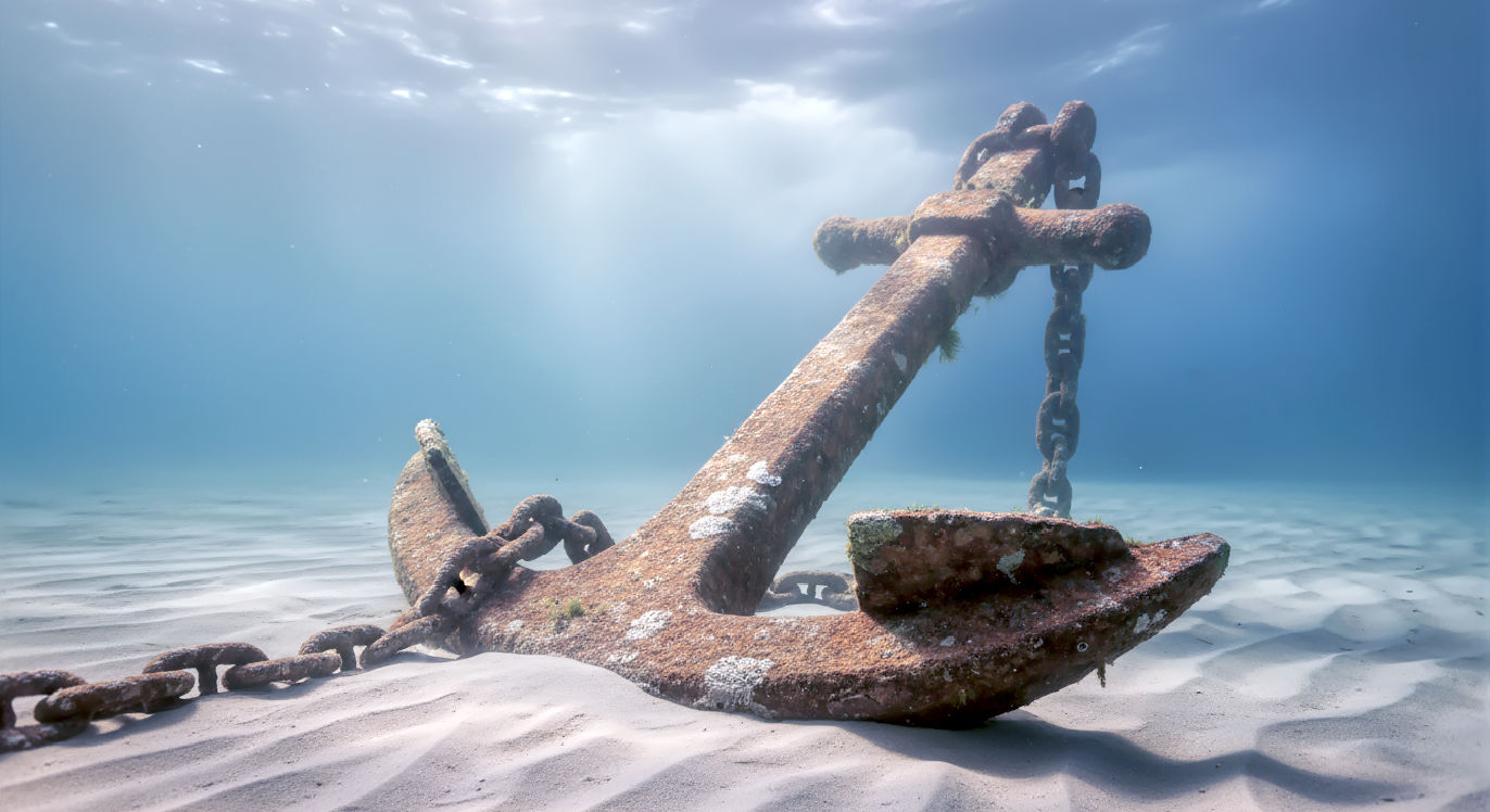 Professional DSLR photo, 16:9, diffused underwater light. Subject: A large, heavy, rusted ship's anchor lodged firmly in the sand on the seabed. Foreground: The massive anchor fluke and part of its thick chain, partially buried, symbolizing the "anchor for the soul." Background: Softly blurred, turbulent water with shafts of light breaking through from the surface, suggesting a storm above. Mood: Secure. Note: NO text, NO abstract graphics. The feeling should be one of profound stillness and stability, even with chaos implied far above.