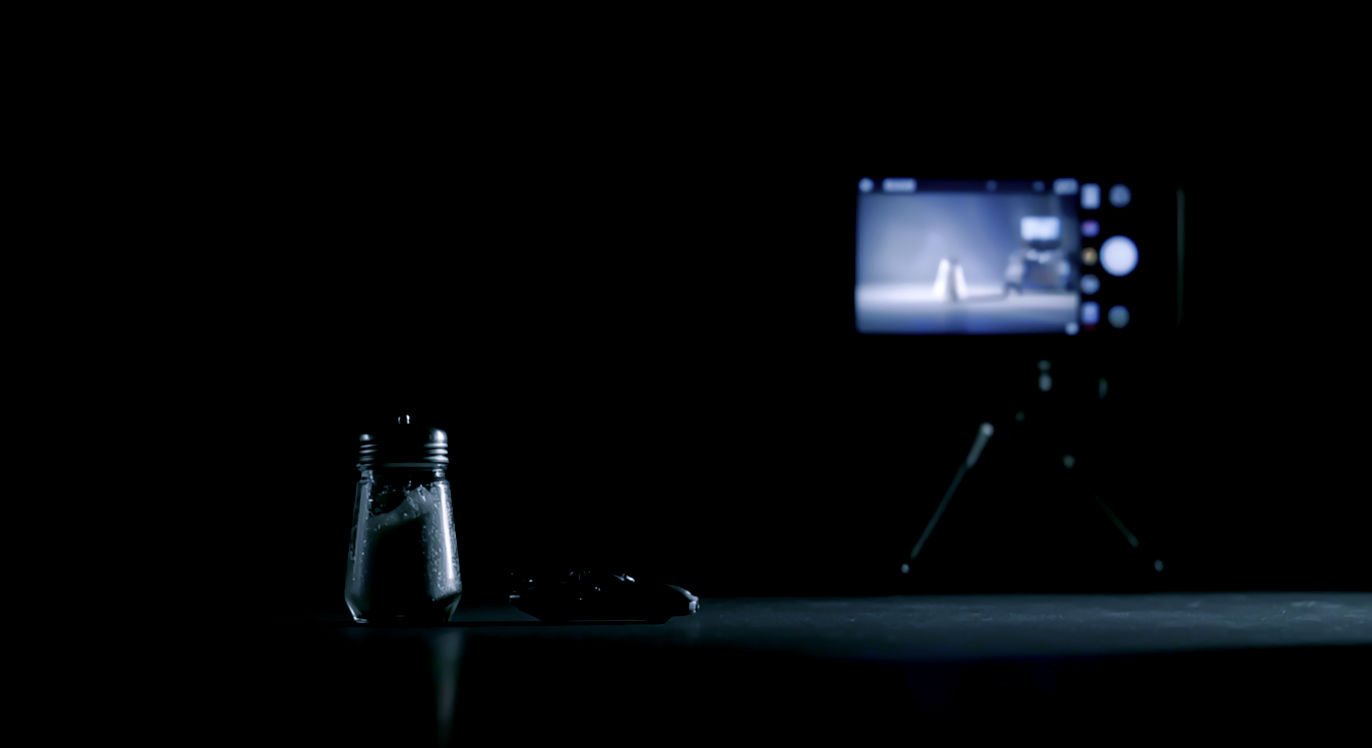 Professional DSLR photo, 16:9, dramatic side light, as if on a tiny "film set." Subject: A single, simple object (like a salt shaker, a toy car, or a coffee mug) on a plain, dark surface. Foreground: The subject is positioned slightly off-center, captured mid-movement. Background: A smartphone on a makeshift "tripod" (e.g., taped to a heavy book or cup) is visible but blurred, pointing at the subject. Mood: Magical. Note: NO text, NO abstract graphics. The image should feel like a "behind-the-scenes" look at the magic.