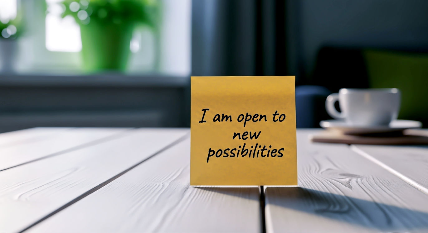 Professional DSLR photo, 16:9, soft morning light. Subject: A single, premium sticky note (perhaps a warm yellow or craft paper brown) with the words "I am open to new possibilities" handwritten in clean, elegant black ink. Foreground: The sticky note is placed on the clean, wooden grain of a desk. Background: A blurred-out, cozy room with a healthy green plant and a coffee mug, suggesting a fresh start to the day. Mood: Hopeful Note: NO text, NO abstract graphics. Avoid people.