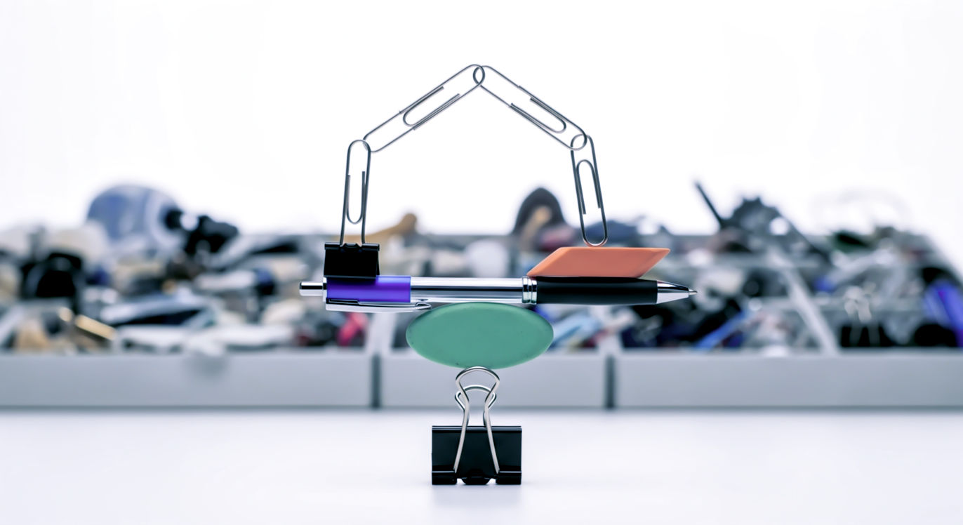 Professional DSLR photo, 16:9, bright, clean studio light. Subject: A small, whimsical sculpture balanced precariously, made of 5-7 common desk items (e.g., a binder clip for a base, a pen as a tower, paperclips linked together, an eraser). Foreground: The sculpture itself, in sharp focus, showing the different textures of plastic, metal, and rubber. Background: A blurred "junk drawer" filled with other random objects, providing context for the "found" items. Mood: Playful. Note: NO text, NO abstract graphics. The focus is on the creative re-use of everyday items.
