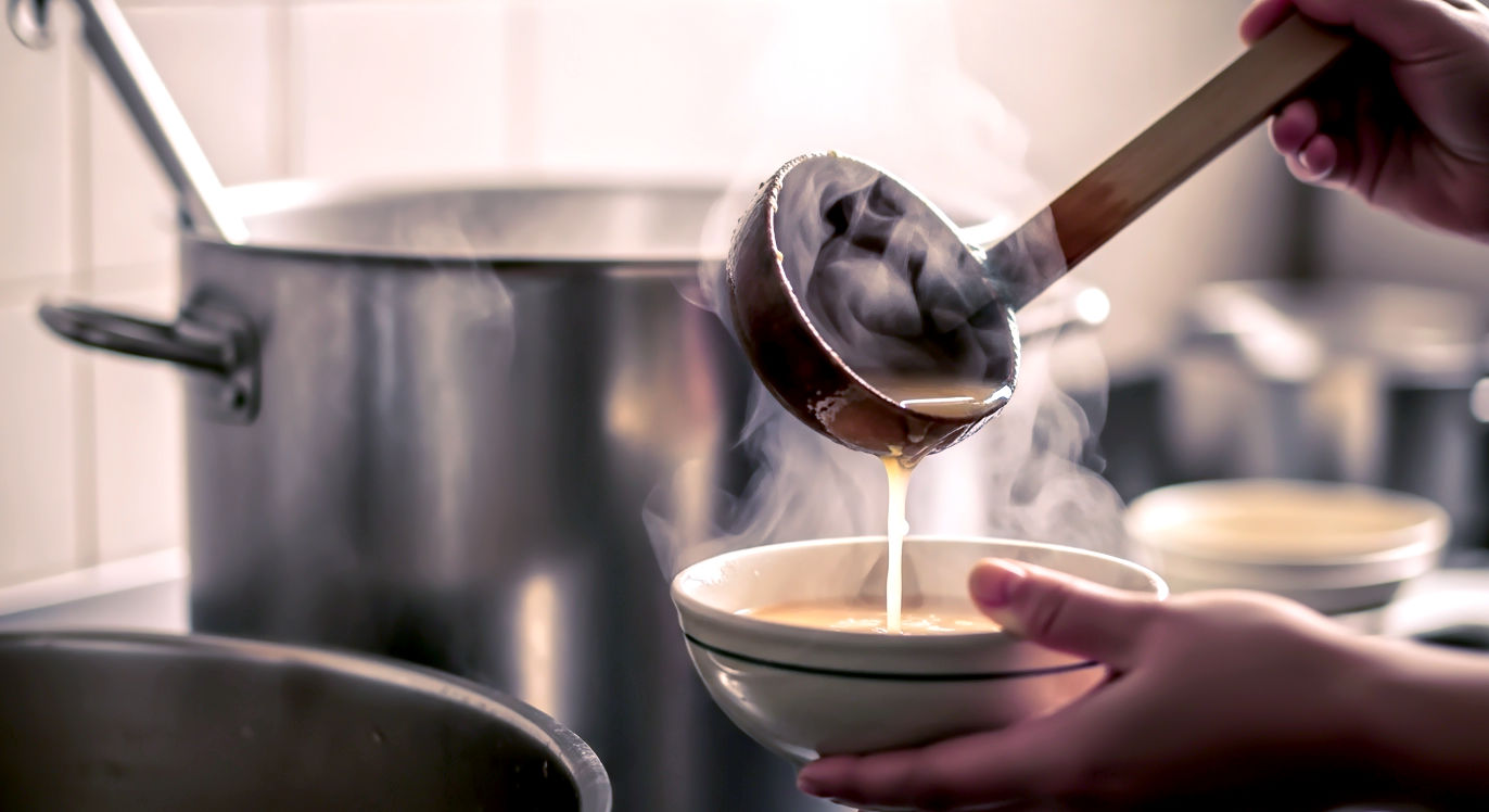 Professional DSLR photo, 16:9, warm, humble indoor light (like a community kitchen). Subject: A close-up shot from a low angle, showing hands holding a simple wooden ladle, serving thick, steam-rising soup. Foreground: The ladle itself, with soup dripping, over a simple ceramic bowl held by unseen hands. Background: A large, slightly blurred stainless steel soup pot, implying a large quantity made for many people. Mood: Generous. Note: NO text, NO abstract graphics. The focus is on the *act* of serving and sharing.