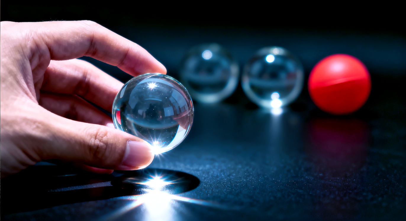 Professional DSLR photo, 16:9, studio side light. Subject: A hand gently holding a single, delicate, clear glass ball. Foreground: The bright, sharp reflection of light on the glass ball's surface. Background: Two other glass balls and one red rubber ball, all blurred, resting on a dark, reflective surface. Mood: Focused 
