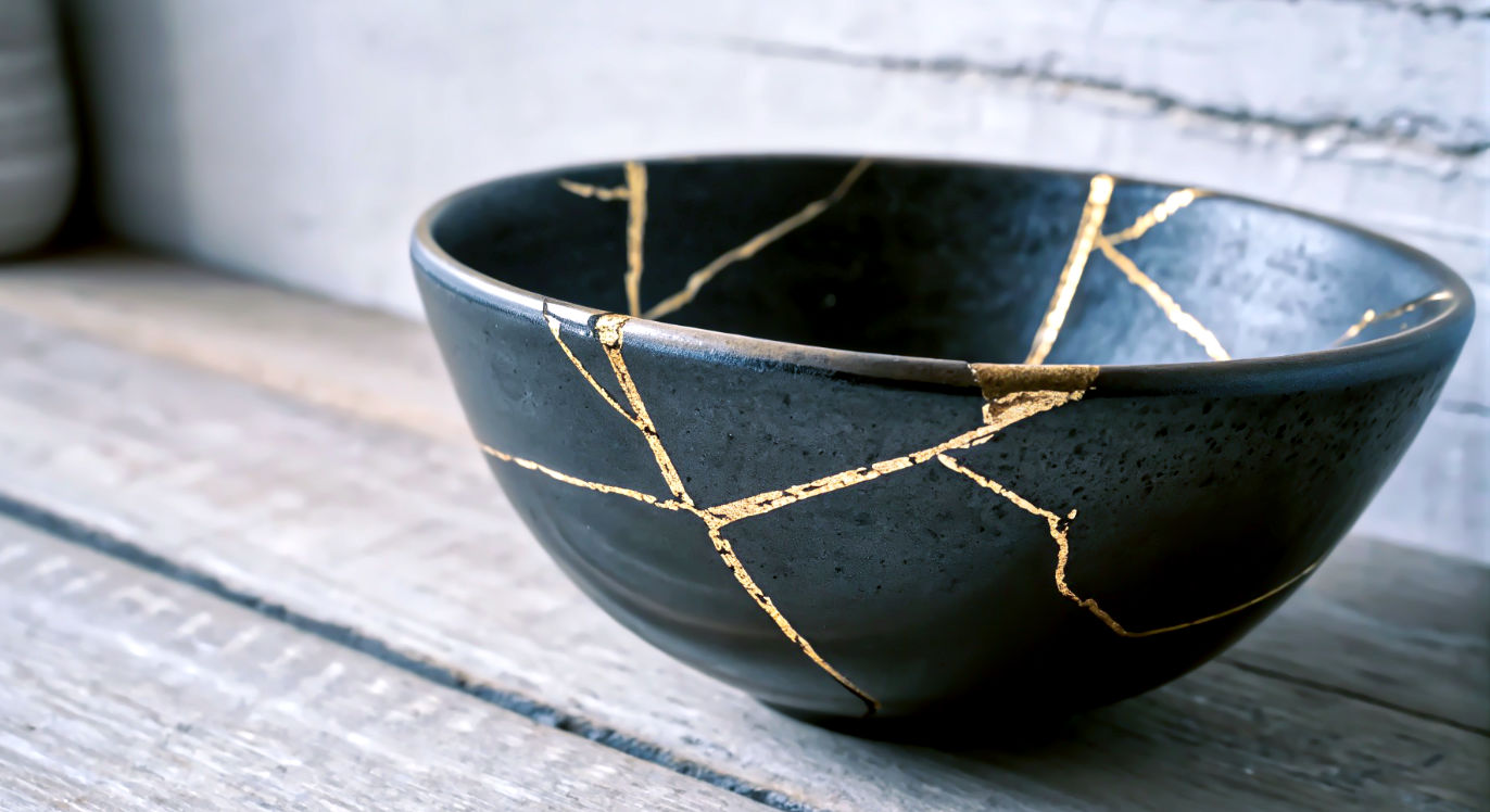 Professional DSLR photo, 16:9, soft side light. Subject: A close-up of a piece of kintsugi (the Japanese art of mending broken pottery with gold). Foreground: A brilliant, textured gold seam running through a piece of simple, dark ceramic. Background: The blurred, simple texture of the rest of the ceramic bowl. Mood: Resilient 