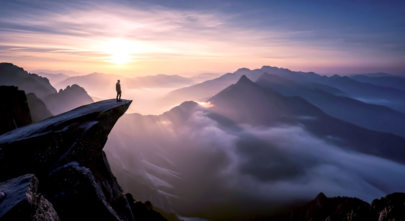 Wide-angle, 16:9. A tiny silhouette of a person on a cliff, looking over a vast, misty mountain range at sunrise. Mood: Awe, Expansive.