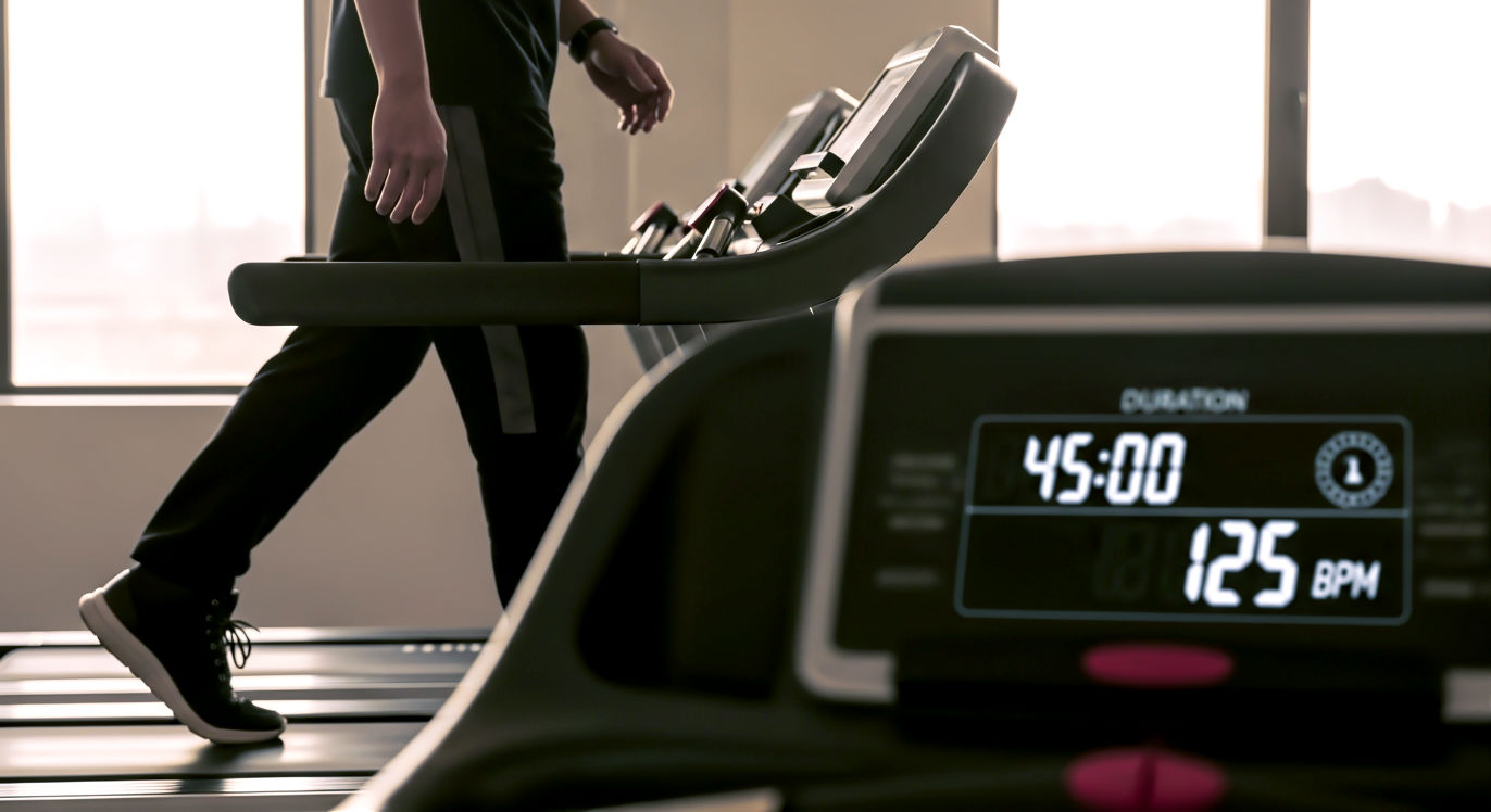 Professional DSLR photo, 16:9, golden-hour side light. Subject: A side-on view of a pair of well-worn trainers on a treadmill, captured mid-stride in a steady walk. Foreground: The treadmill console in soft focus, showing a long duration (e.g., "45:00") and a steady heart rate (e.g., "125 BPM"). Background: The calm, empty gym environment, with the warm light streaming through a large window. Mood: Steady