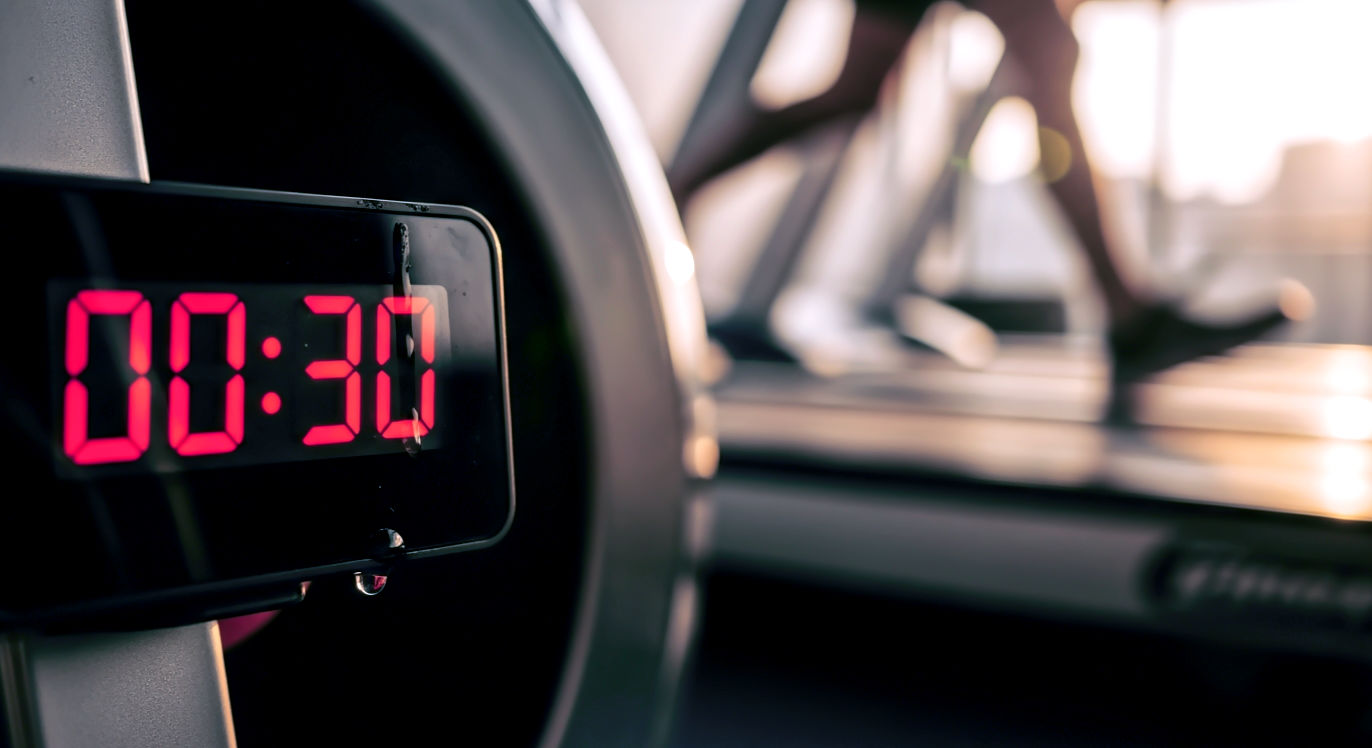 Professional DSLR photo, 16:9, golden-hour side light. Subject: A close-up of a digital gym timer's red numbers, sharply in focus, showing "00:30". Foreground: A single bead of sweat dripping down the timer's casing or a blurred hand reaching past to slam a button. Background: The blurred, kinetic motion of a spinning bike's flywheel or a person's legs sprinting on a treadmill. Mood: Intense
