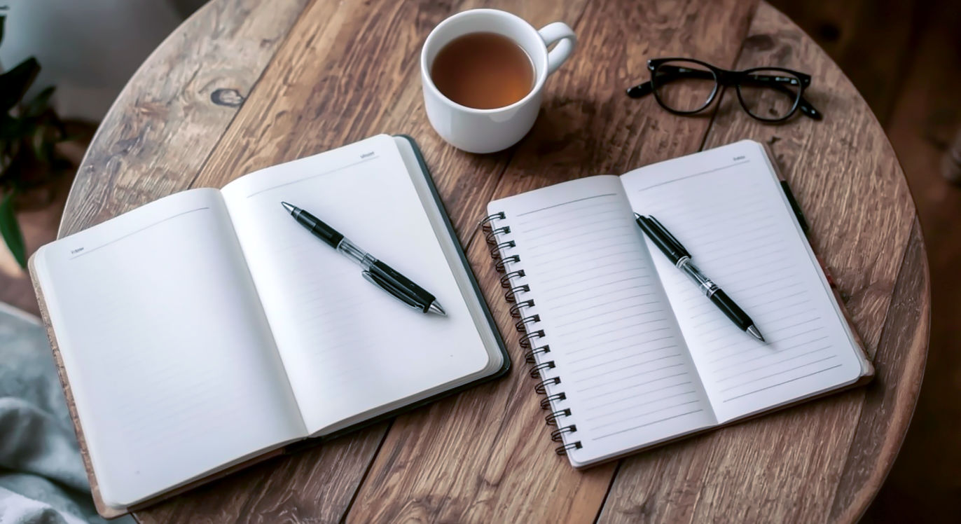 A warm, inviting photograph showcasing a hybrid journaling approach. On a cozy, slightly rustic wooden table, an open guided journal lies next to a separate, open blank notebook. Both have pens resting on their pages, suggesting active use. Perhaps a comforting cup of tea or coffee, and a pair of reading glasses are nearby. The composition should convey flexibility, balance, and personalization, suggesting that different tools can coexist harmoniously to support a holistic practice. Use soft, ambient lighting and a shallow depth of field to create a sense of comfort and reflection.