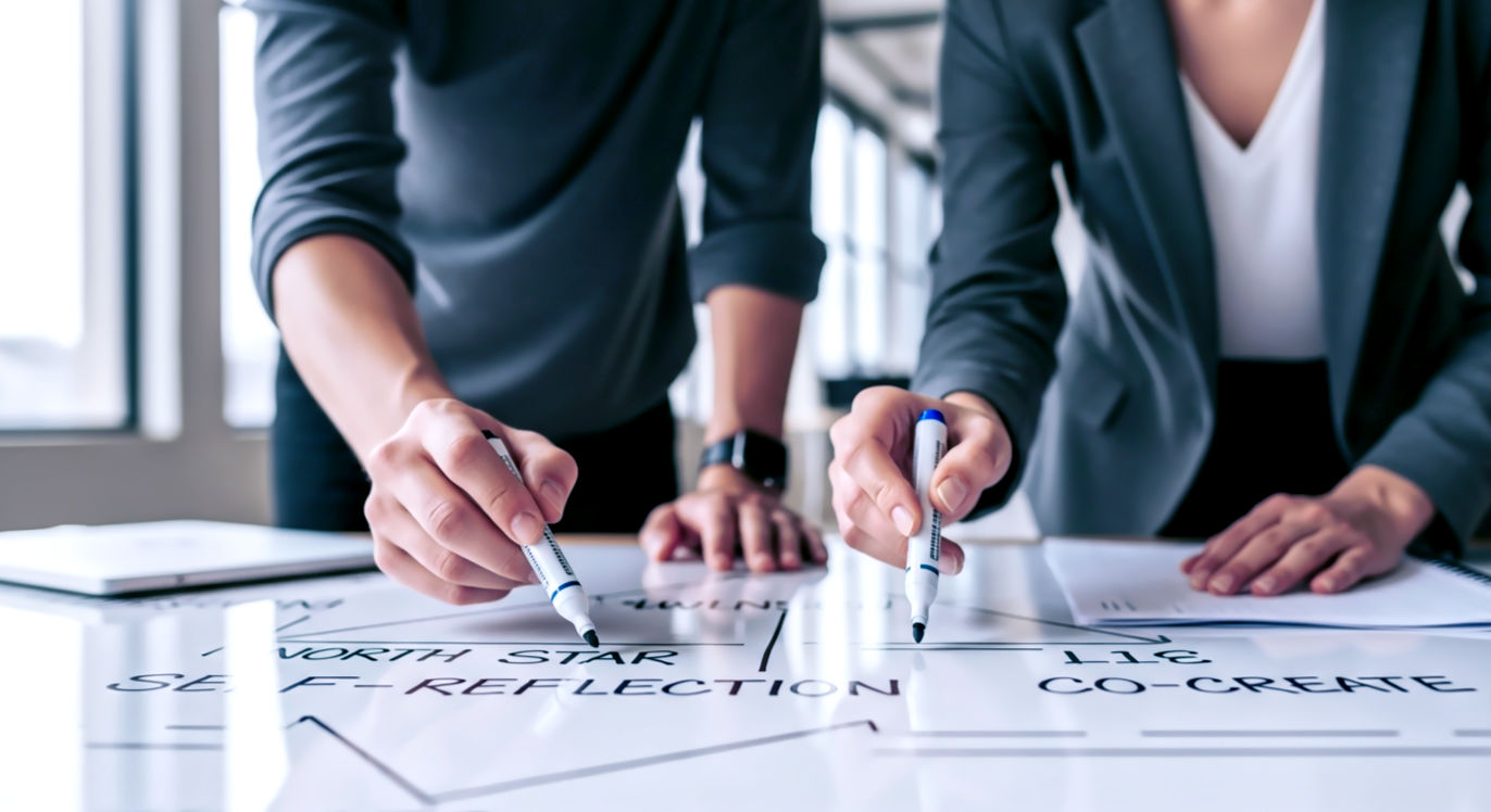Professional DSLR photo, 16:9, bright, inviting natural light. Subject: Two people (diverse, professional attire, one appearing as a manager and one as an employee) actively collaborating, leaning over a large whiteboard. The whiteboard has a few key phrases like "North Star," "Self-Reflection," and "Co-Create" written on it. They are both holding markers. Foreground: Their hands, holding markers, pointing to the whiteboard. Background: A modern, open-plan office space, slightly blurred. Mood: "Collaborative"