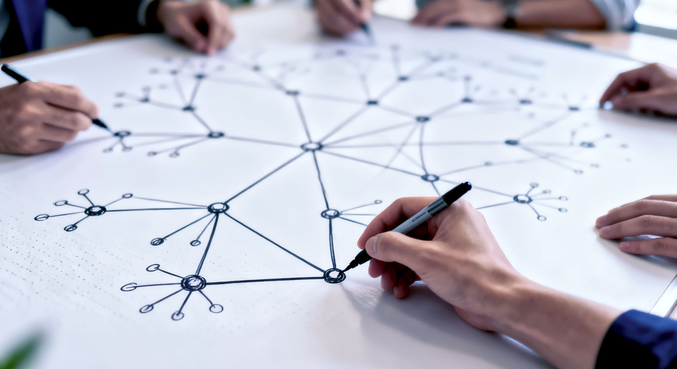 Professional DSLR photo, 16:9, clean indoor light. Subject: A close-up shot of several hands collaboratively drawing a complex, organic diagram on a large whiteboard. The diagram shows small nodes connecting and growing into larger, interconnected networks, like a blueprint for a collective system. Foreground: One hand drawing a connecting line between two nodes. Background: The rest of the sprawling diagram, slightly out of focus, implying scale and future planning. Mood: Collaborative Note: NO text. This image symbolizes the 'design' of future growth, collective effort, and scaling up from individual mindset to organizational culture.
