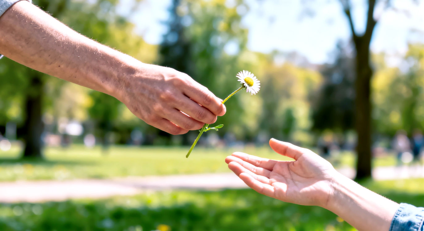 A vibrant, professional, lifelike photograph capturing the "ripple effect" of kindness. The composition is a close-up focusing on three different people's hands, one after another in a chain. The first hand is passing a single, simple wildflower to a second hand, which is in the process of receiving it. The second hand is already angled to pass the flower to a third hand, which is just entering the frame. The background is a soft-focus, out-of-focus blur of a bright, outdoor community setting, like a park. The lighting is warm and natural, suggesting a beautiful day. The mood is one of connection, community, and the simple, quiet spread of a good deed.