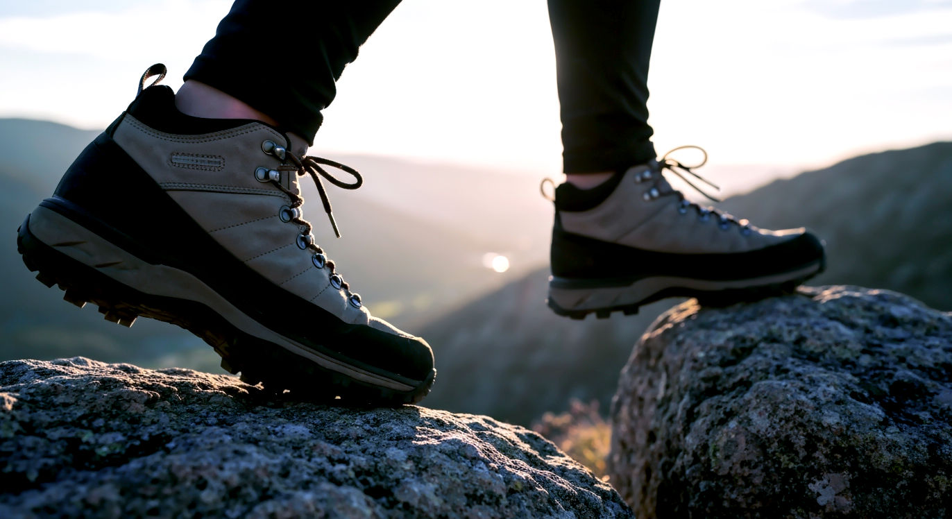 Professional DSLR photo, 16:9, golden-hour side light. Subject: A hiker's boot, in sharp focus, planted firmly on one rock, with the other foot blurred in motion, stepping across a small-but-clear gap to the next rock. Foreground: The texture of the rock and the sole of the boot, showing grip and action. Background: A blurred, beautiful, and expansive natural vista (e.g., a valley or mountain range) suggesting a forward-looking journey. Mood: Aspirational. Note: NO text, NO abstract graphics. Focus on the action of "leaping" or "stepping up."