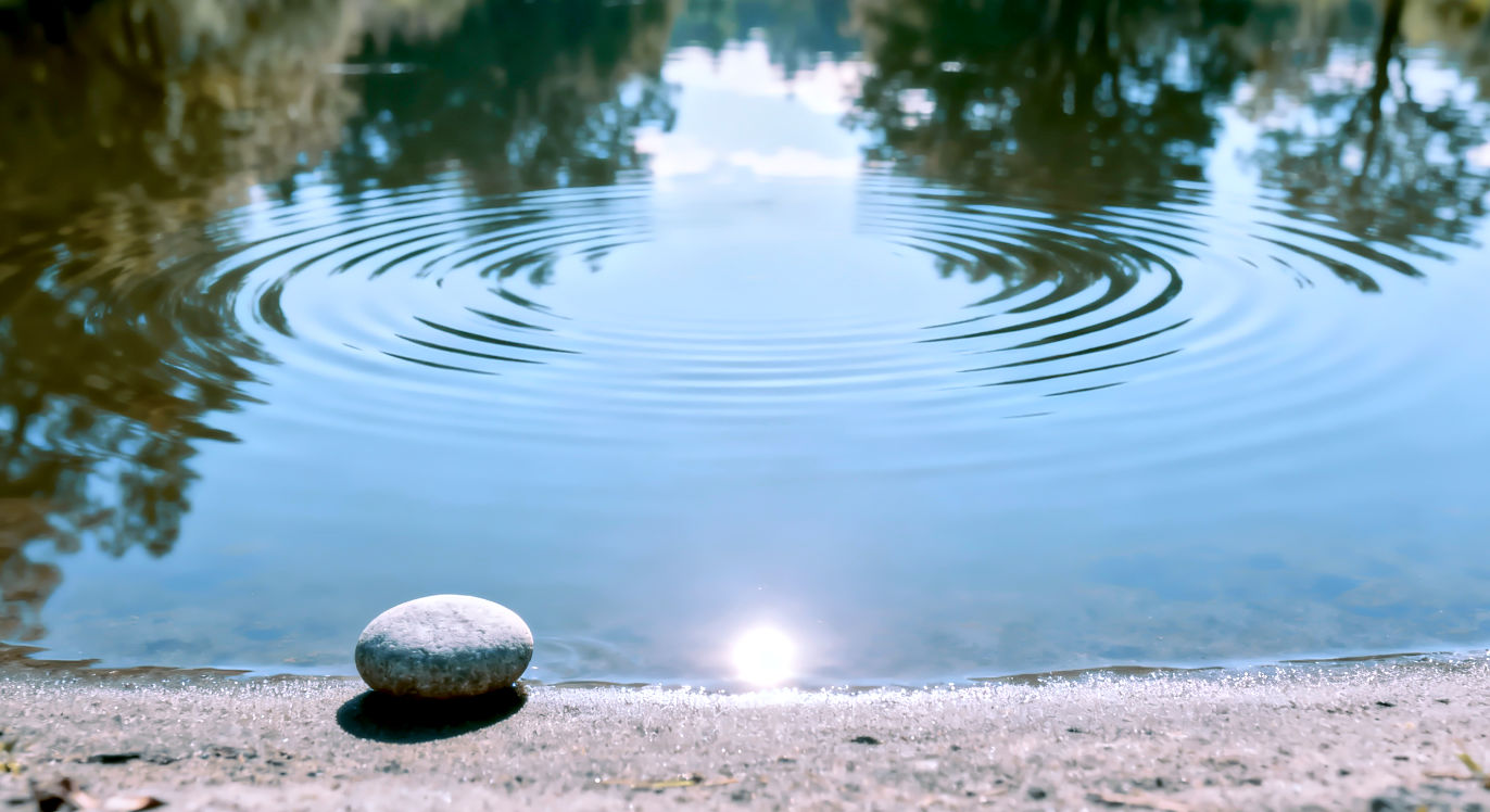 > Professional DSLR photo, 16:9, bright, even, ambient light. > **Subject**: A serene, still pond or lake surface with very subtle, perfectly symmetrical ripples spreading outwards from a central, unseen point. > **Foreground**: A single, smooth, perfectly rounded pebble at the edge of the water, catching a highlight. > **Background**: A softly blurred, harmonious natural landscape (trees, sky) reflecting in the water. > **Mood**: Harmonious > **Note**: NO text, NO abstract graphics. The image should symbolize perfect rhythm and stability.