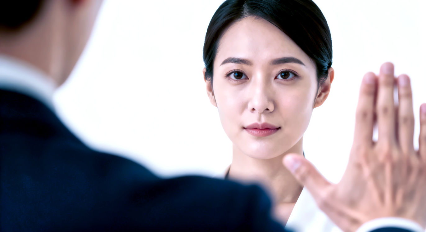 A powerful, tight-shot professional photograph focusing on a person's calm and composed face as they hold a boundary. They are looking directly at the viewer (or just off-camera) with a neutral, unshakeable, and polite expression—not angry, just firm. In the foreground, another person's hand is visible and slightly out of focus, gesturing insistently. The main subject is not reacting to the pressure, remaining perfectly still and centered. The lighting is clean and bright, isolating the subject. The mood is one of quiet confidence, resilience, and unshakeable (but polite) firmness.
